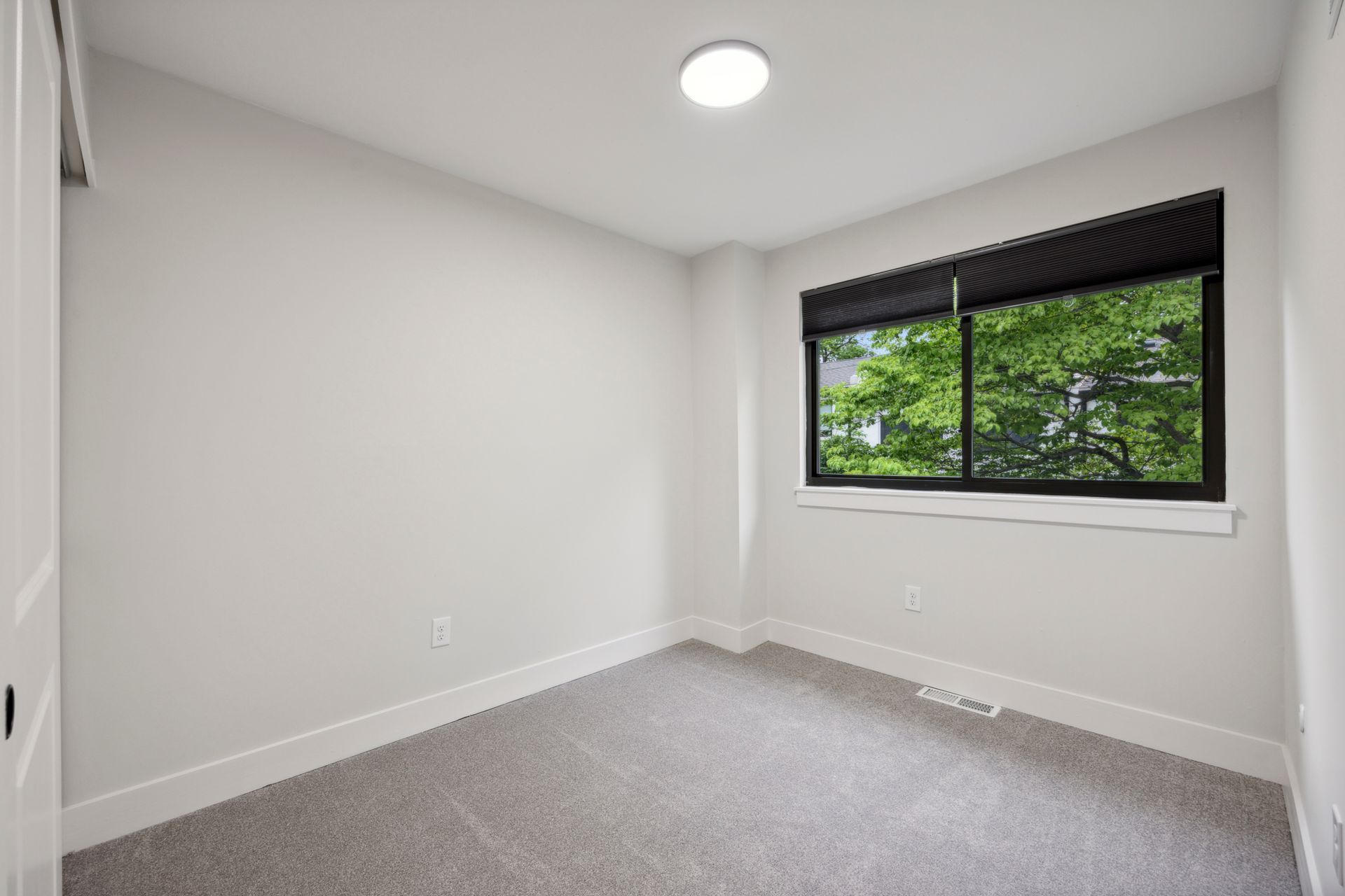 Empty bedroom with gray carpet, white walls, a window with a dark shade, and a circular ceiling light.
