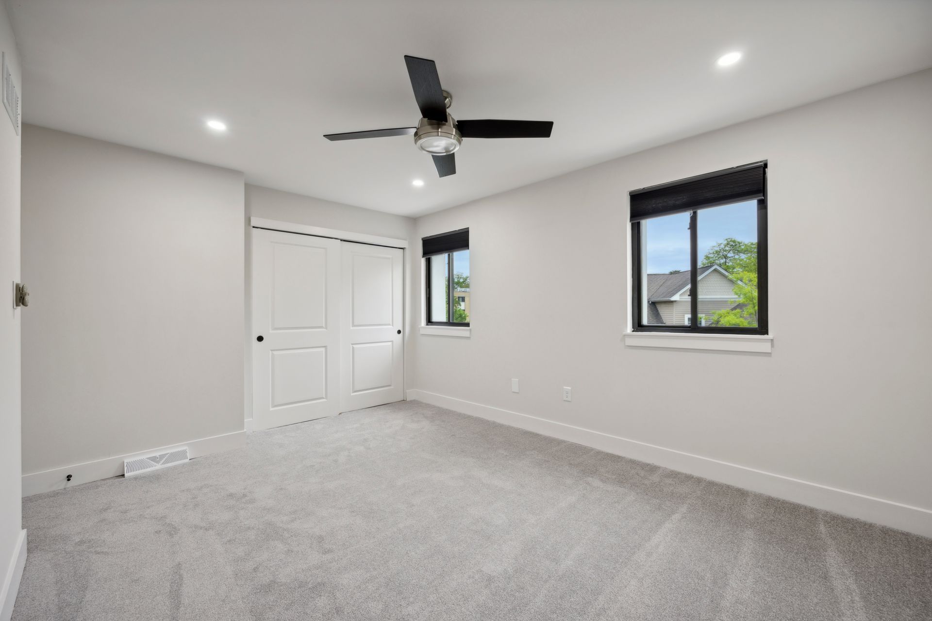 Empty bedroom with gray carpet, white walls, two windows, and a ceiling fan.