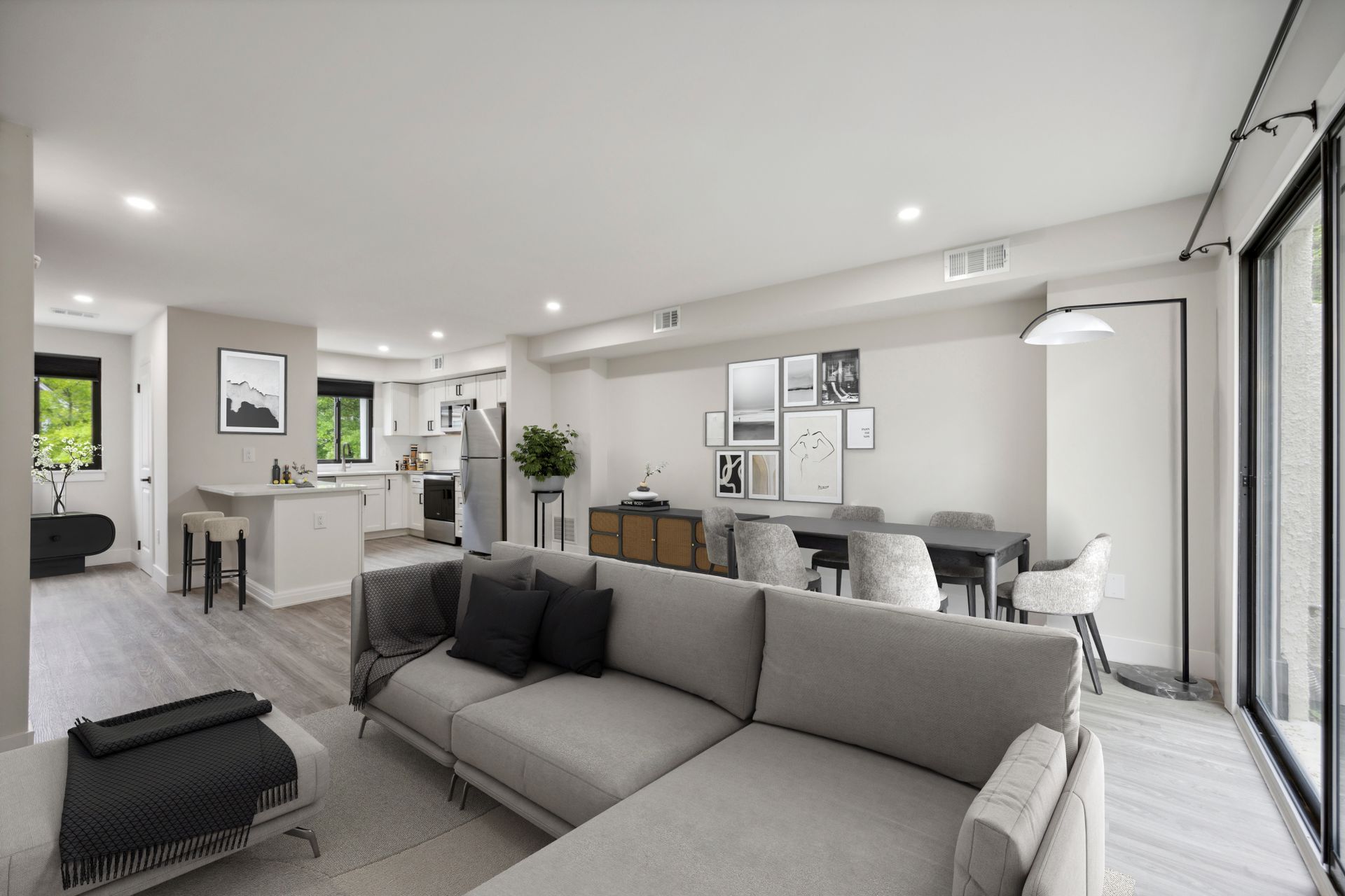 Open-concept living space with a gray sectional sofa, dining area, and kitchen. Light wood floors and neutral walls.