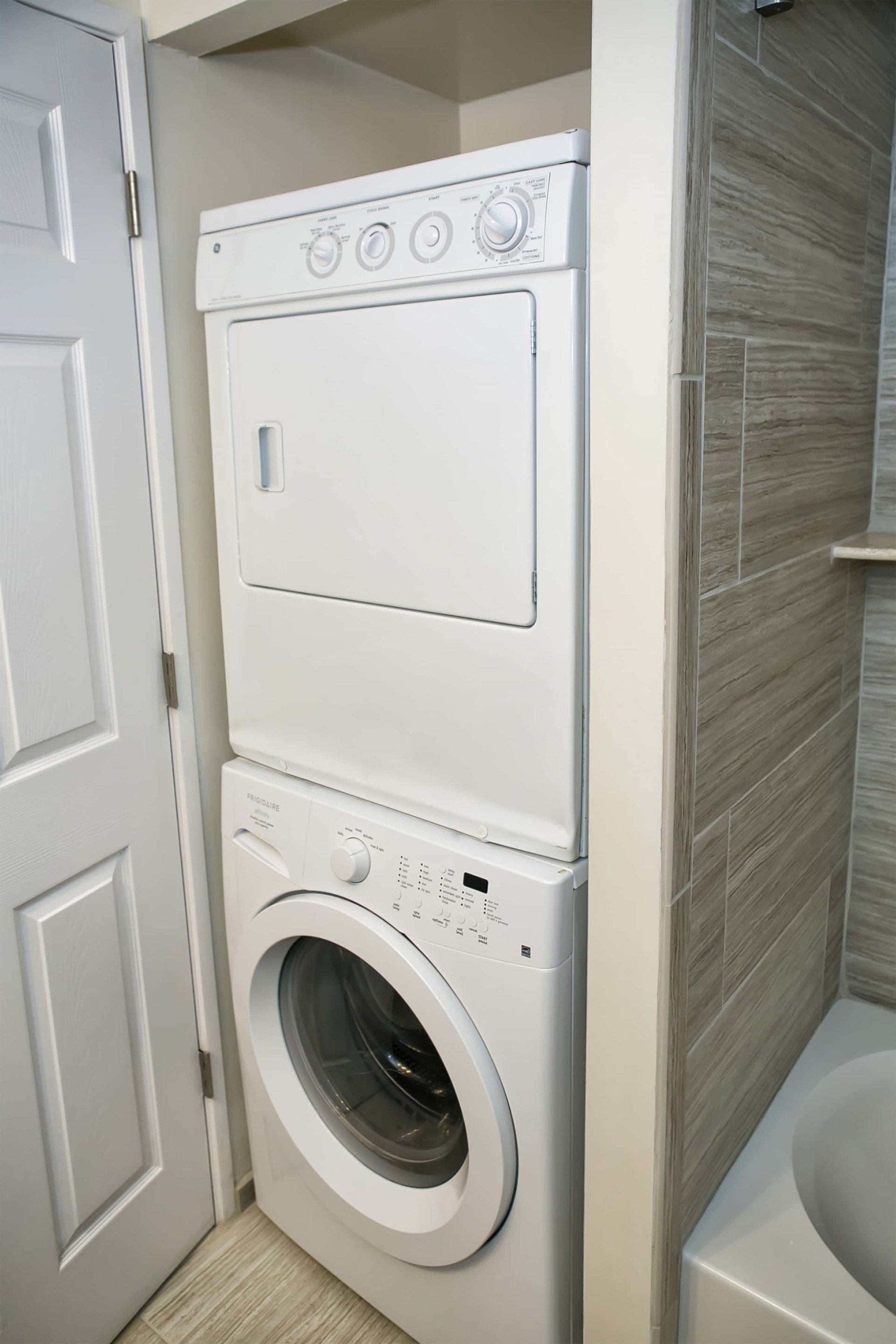 Stacked white washer and dryer in a small alcove next to a bathroom door and tiled wall.