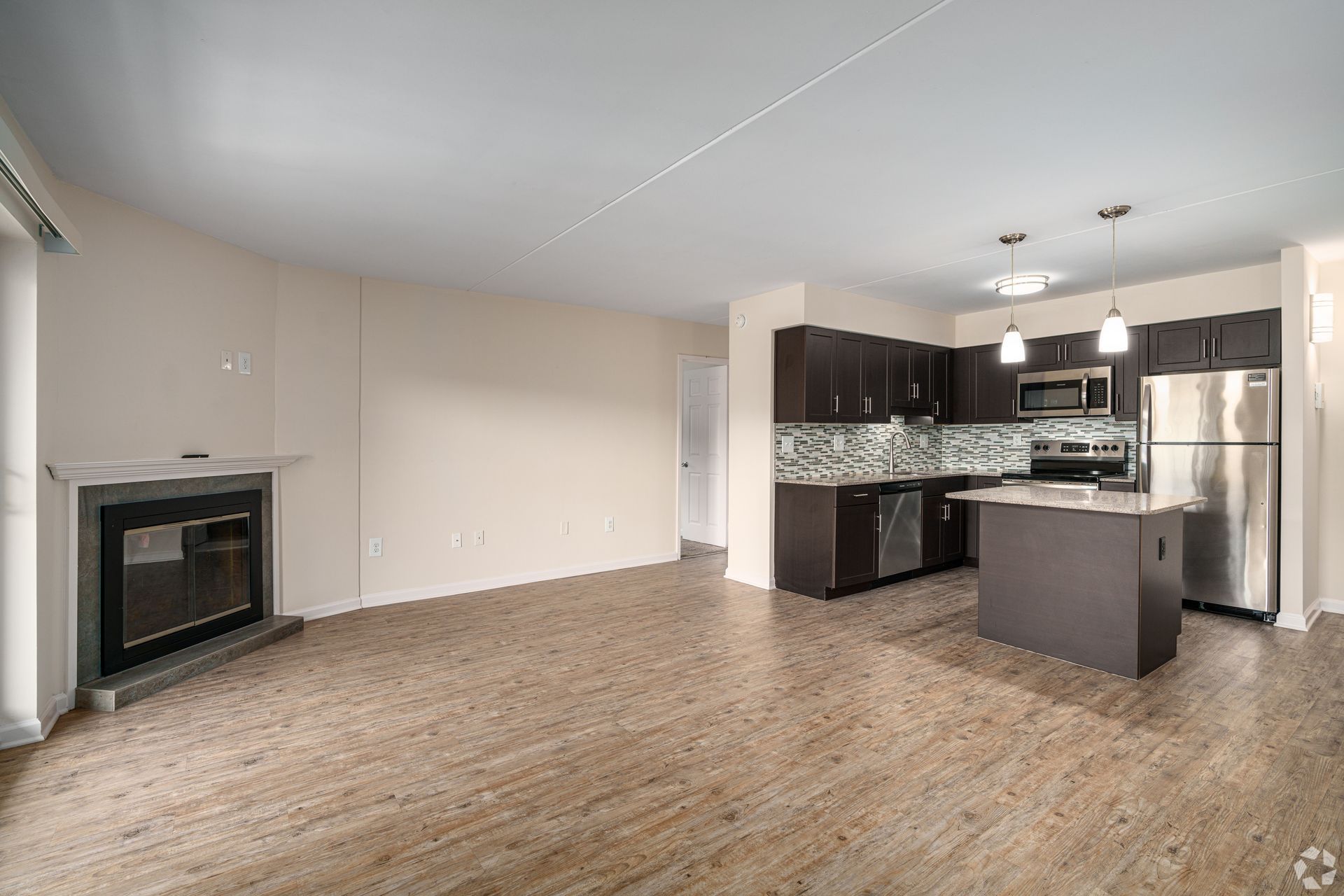 Empty apartment interior with kitchen, fireplace, and wood-look flooring.