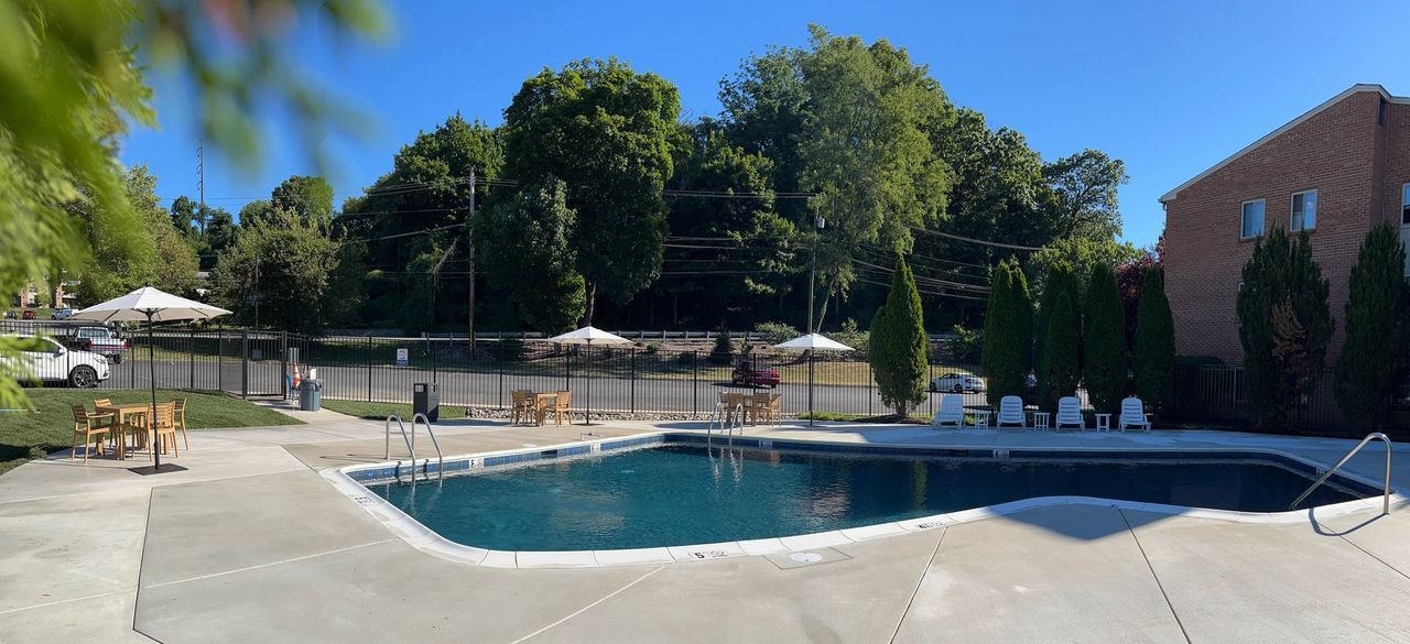 A pool surrounded by concrete, trees, a fence, and a brick building, under a blue sky.