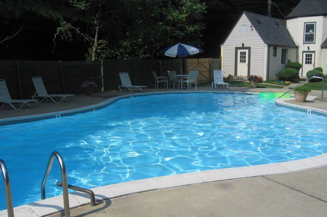 Pool with blue water, white lounge chairs, umbrella, and a small building in the background.