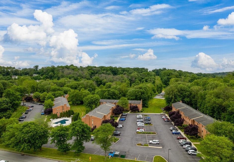 Aerial view of apartment complex with parking, pool, and surrounding trees under a blue sky with clouds.
