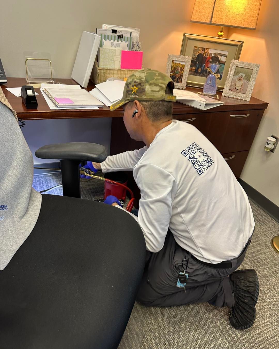 Person kneels to work under a desk in an office setting. They wear a white shirt, camo hat, and work pants.