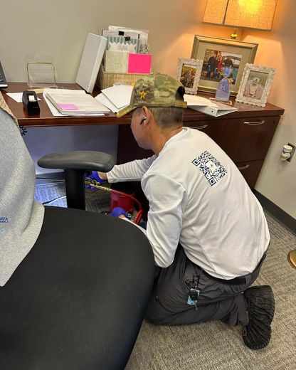 Person kneels to work under a desk in an office setting. They wear a white shirt, camo hat, and work pants.