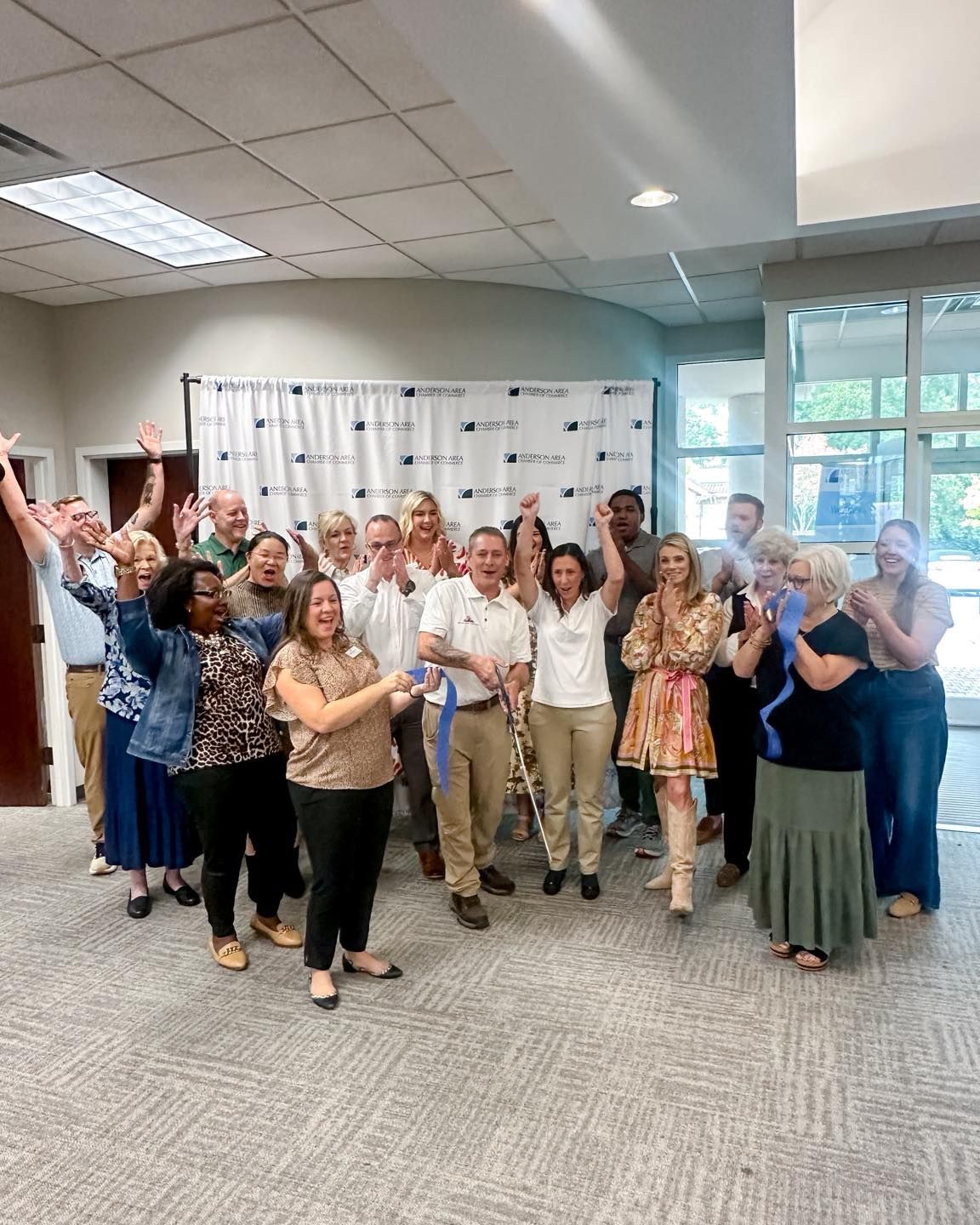 Group of people celebrating, some waving arms. Ribbon cutting in a light-filled space.