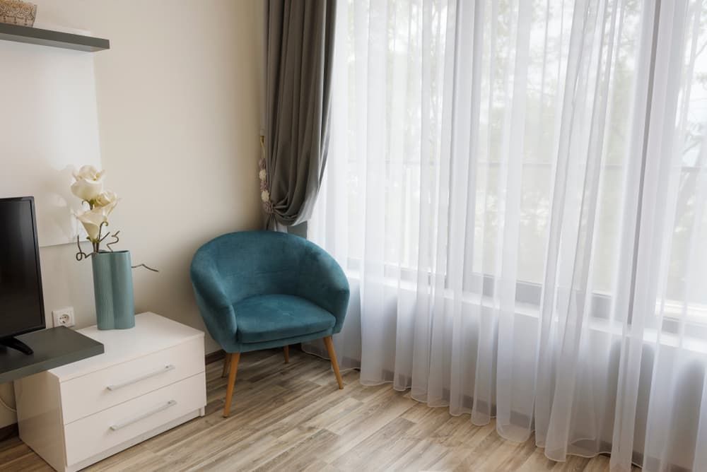 A Teal Armchair Sits by A Window with Sheer Curtains Next to A White Nightstand — U Curtains in Acacia Ridge, QLD