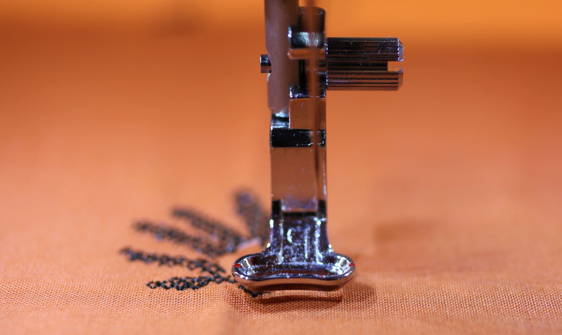 Prensatelas para máquina de coser sobre un diseño floral bordado en tela naranja. El prensatelas es plateado y brillante.