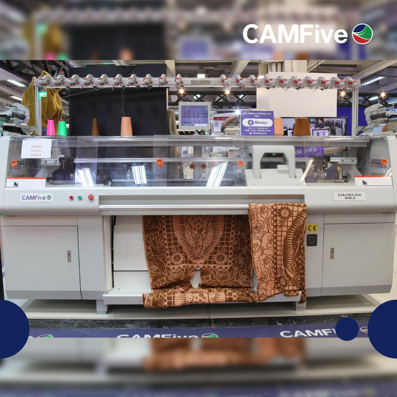 Una gran máquina de tejer industrial en una fábrica, que produce telas estampadas. La máquina es de color gris claro, con una cubierta protectora transparente y rollos de tela adheridos.