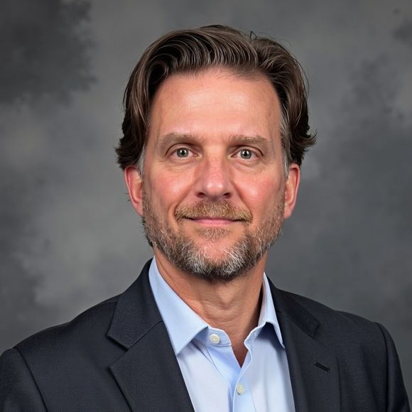 Man in a blazer and blue shirt, smiling, with a short beard, against a dark gray background.