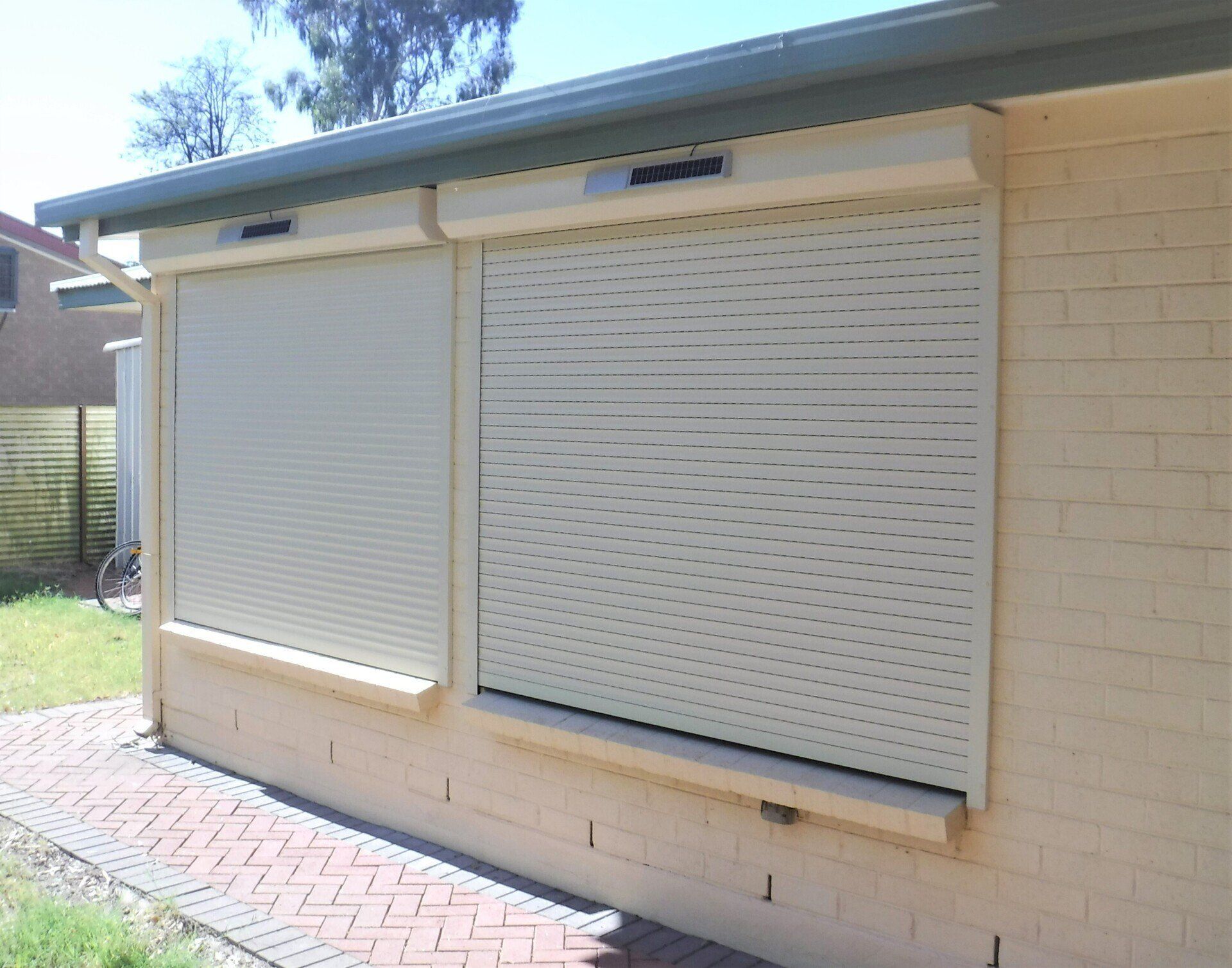 Beige Roller Shutters Over Windows on A Light Yellow Brick Wall — Alice Mobile Blinds in Ciccone, NT