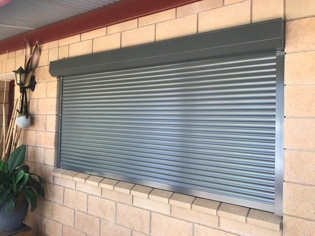 Grey Metal Rolling Shutter Over a Window in A Brick Wall — Alice Mobile Blinds in Ciccone, NT