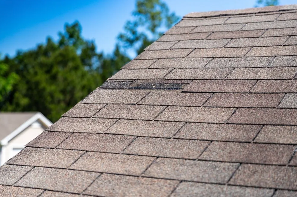 Close-up of a weathered brown shingle roof with a damaged section against a blue sky and green trees.