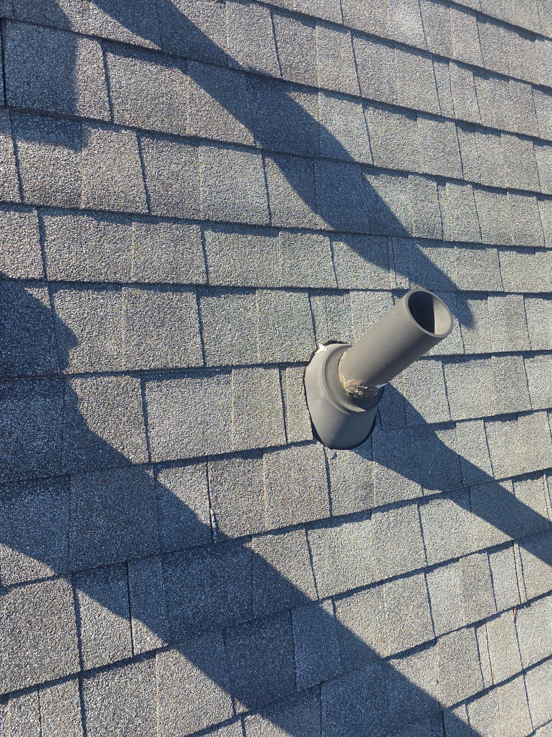 Gray roof shingles with a gray vent pipe extending upwards; shadow of a tall object across the shingles.