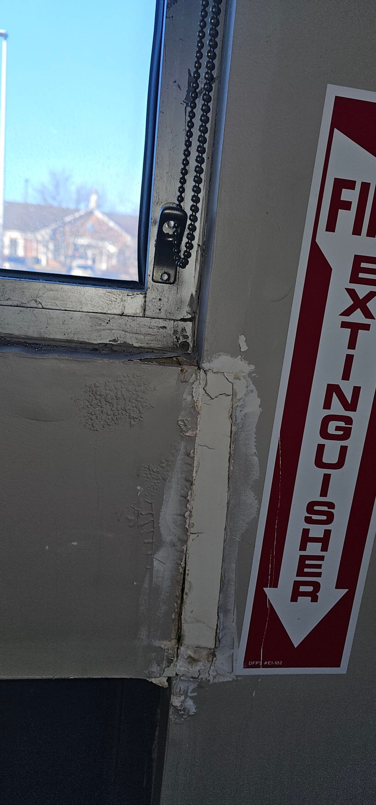 A fire extinguisher sign next to a window, with a view of a building and blue sky outside.