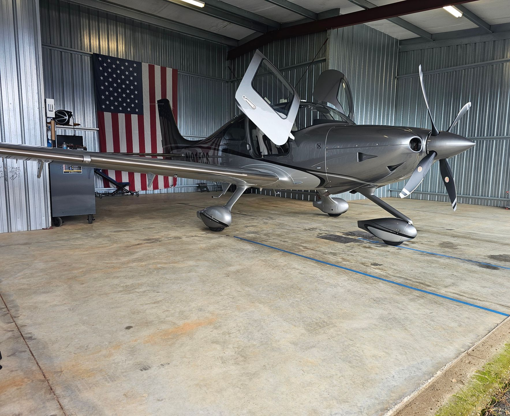 Silver airplane inside a hangar, American flag in background, doors open.