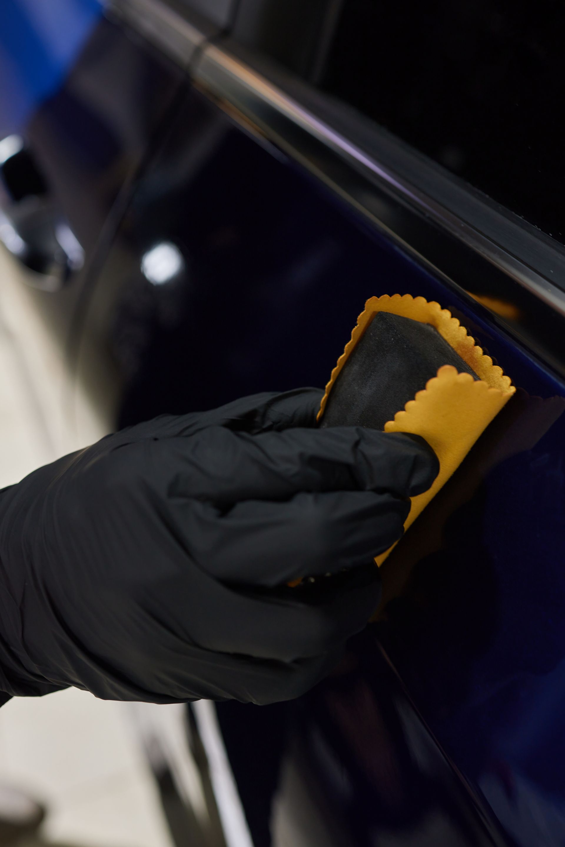 A person wearing black gloves is polishing a blue car with a yellow sponge.