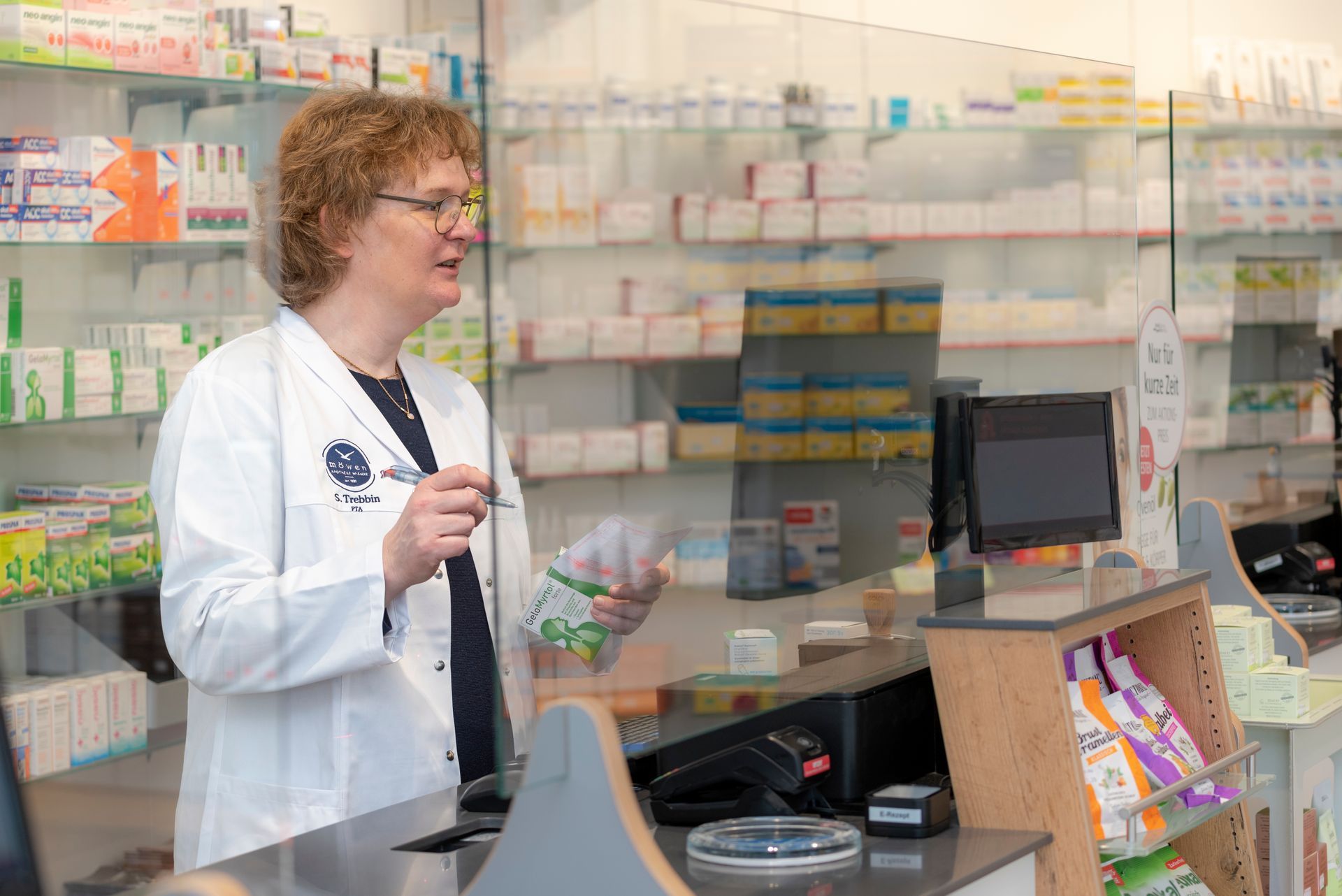 Eine Apothekerin steht hinter einer Glasbarriere in einer Apotheke.