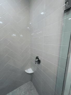 Tiled shower stall with a corner shelf, shower handle, and glass door edge