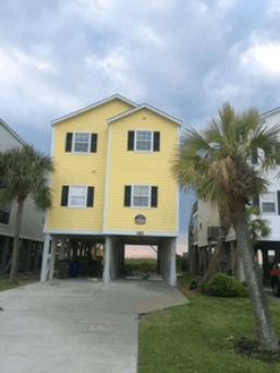 Yellow elevated beach house with a driveway, palm trees, and a cloudy sky