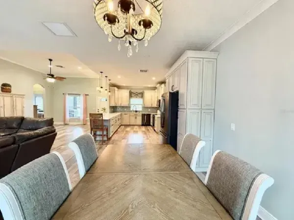Bright open dining and kitchen area with a long wooden table, white cabinets, and a chandelier