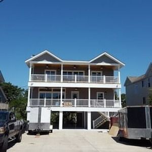 Two-story beach house with white balconies and blue roof, seen from a driveway under a clear sky
