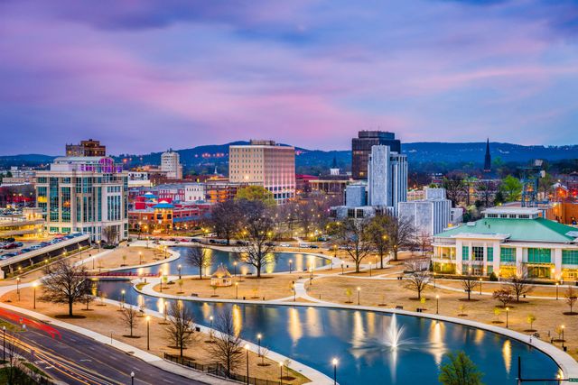City skyline at dusk with a lit waterfront park, bridge, and calm reflective water under a pink-purple sky