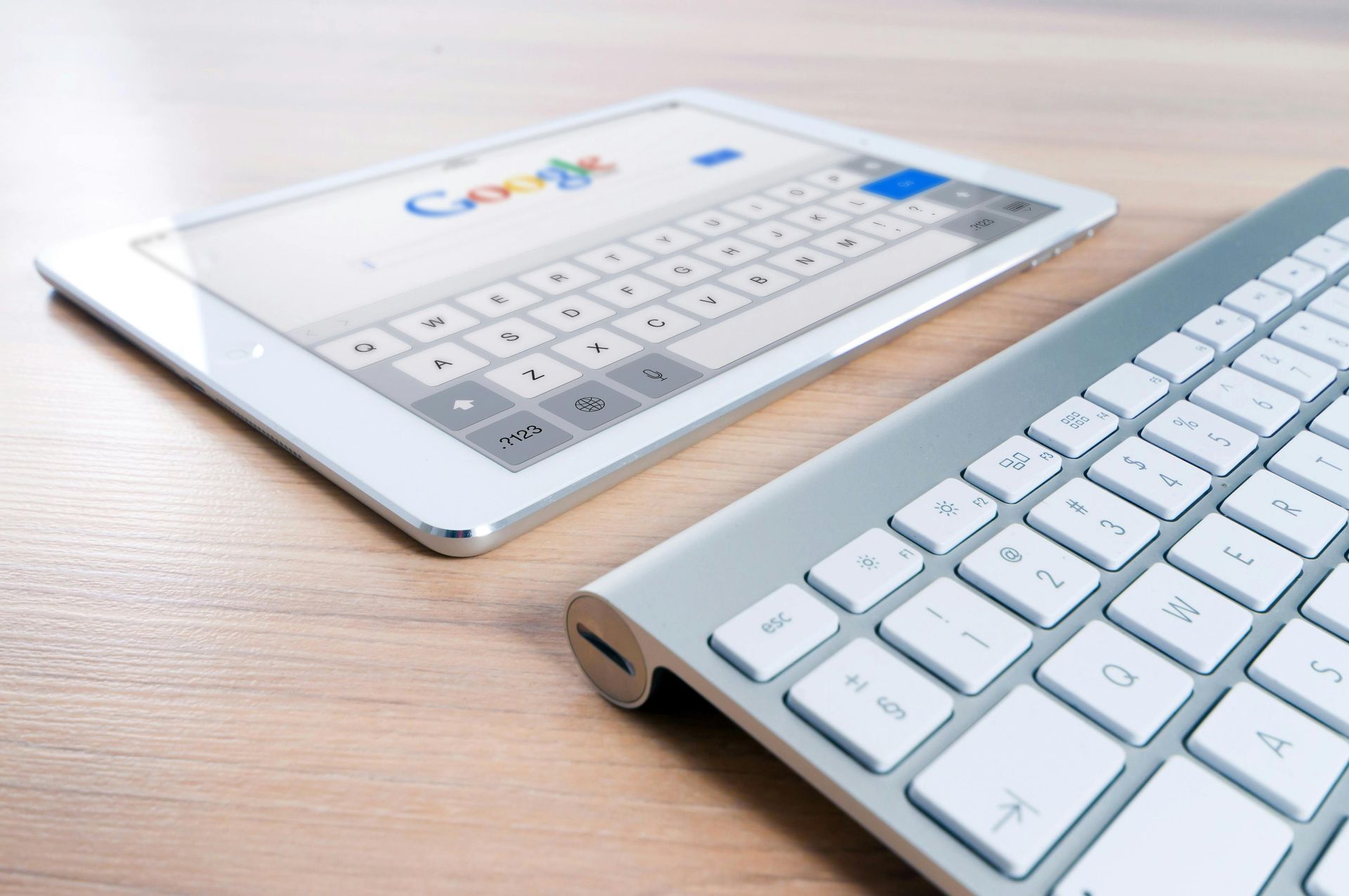 White tablet displaying Google search page next to silver keyboard on a wooden desk.