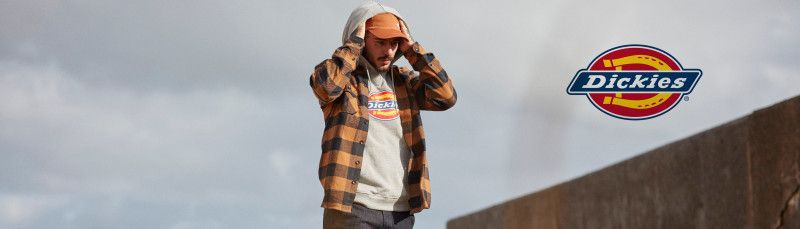 A man wearing a plaid shirt and a hoodie is standing next to a wall.