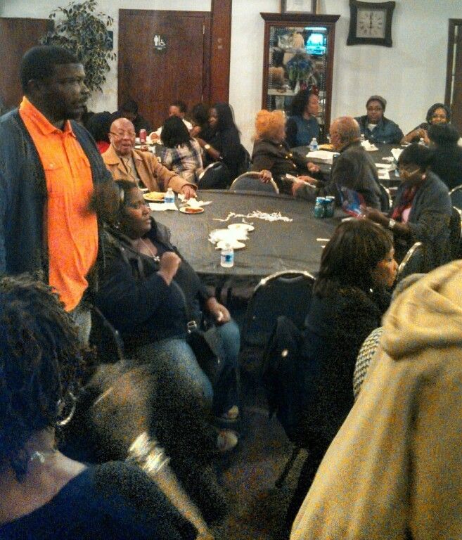 A man in an orange shirt stands in front of a group of people sitting at tables