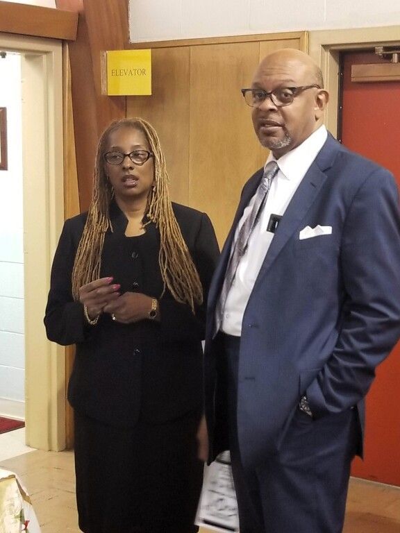 A man in a suit and tie stands next to a woman in a black suit