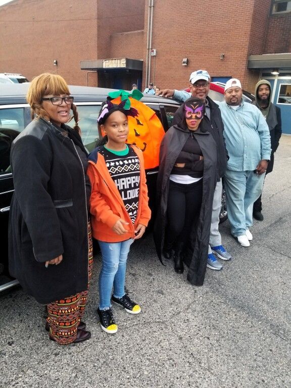 A group of people standing in front of a car with a girl wearing a shirt that says happy halloween