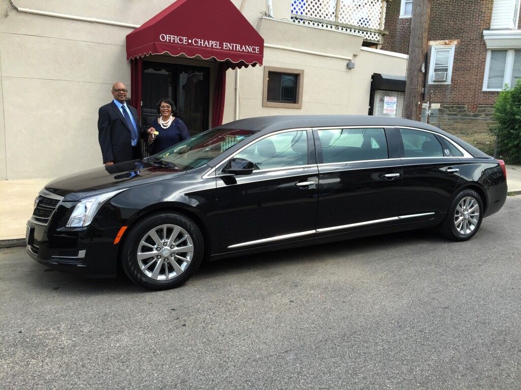 A man and a woman are standing next to a black limousine parked in front of a building.