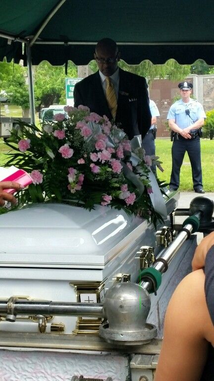 A man in a suit and tie stands behind a white coffin with pink flowers