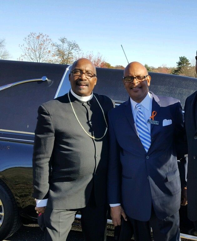 Two men in suits and ties are posing for a picture in front of a black limousine
