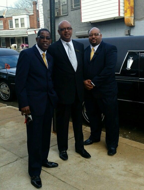 Three men in suits and ties stand in front of a black limousine