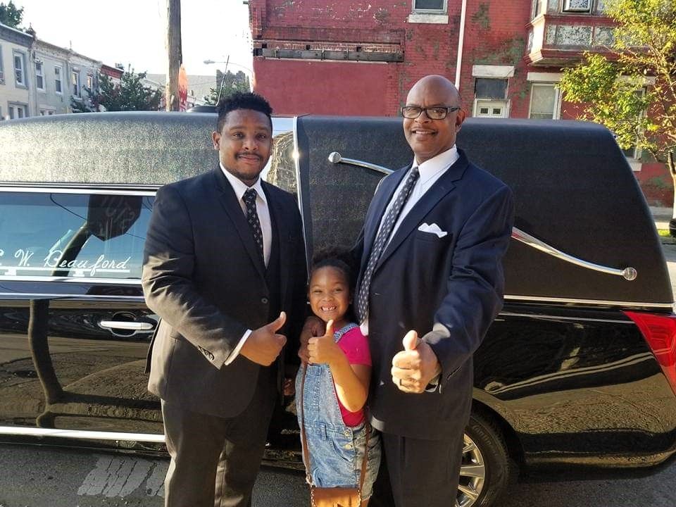 Two men and a little girl are posing in front of a black limousine