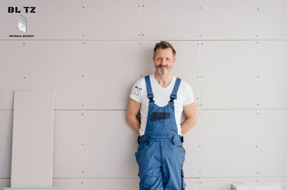 A smiling drywall contractor in blue overalls stands against freshly installed drywall panels with visible seams. This drywall contractor is servicing Essex Rd, Danbury Rd, Kearny, AZ.