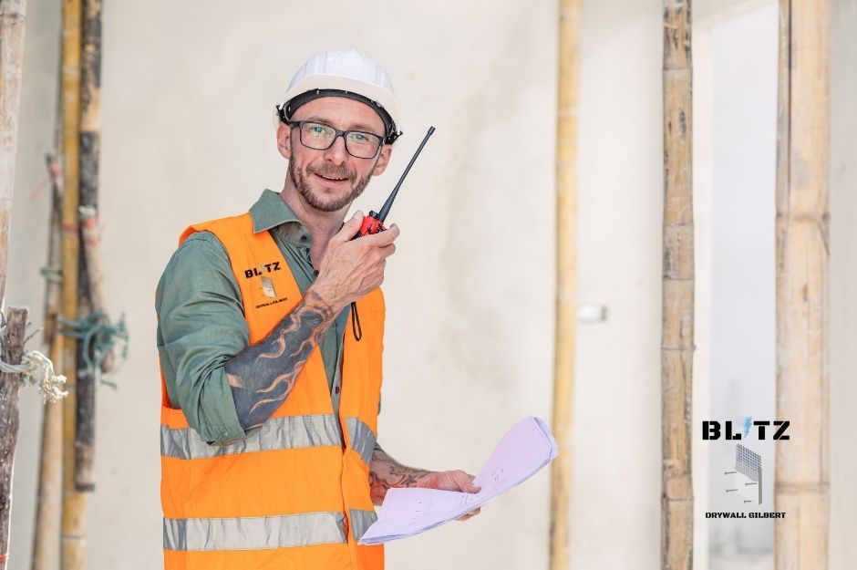 A drywall contractor wearing a white hard hat and an orange safety vest, holding a walkie-talkie and blueprints. He is working on a project for a drywall company near S Stewart and W Vine Ave, Mesa, AZ.