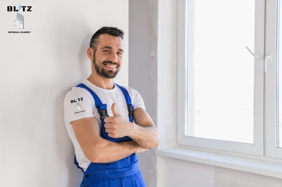 A smiling drywall contractor in blue overalls gives thumbs up in a bright room with white walls and windows. This drywall contractor is servicing W La Osa St, N Chaparral St Oracle, AZ.