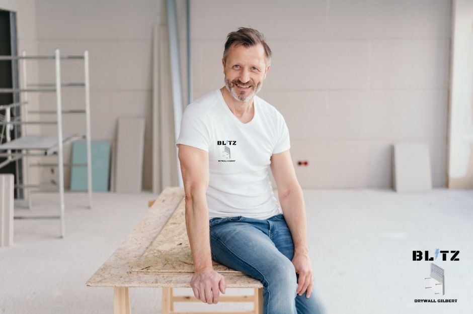 A smiling drywall contractor sits on wooden beam during drywall project with ladder and white walls. This drywall contractor is servicing W Webb Dr, N Douglas Ave, San Manuel, AZ.