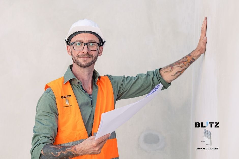 A drywall contractor wearing a white hard hat, an orange safety vest, and glasses stands with one hand resting against the wall. He is working on a project near E Encanto Dr, and E Vista Del Cerro Dr, Tempe, AZ.
