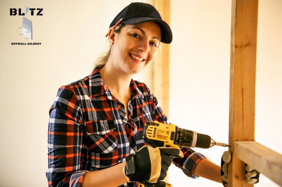 A drywall repair company professional in a plaid shirt and black cap uses a power drill on a residential construction project. She wears work gloves and smiles while working on wooden framing in a renovation space with white drywall walls near E Desert Trumpet Rd, and S 44th St in Ahwatukee Foothills, AZ.