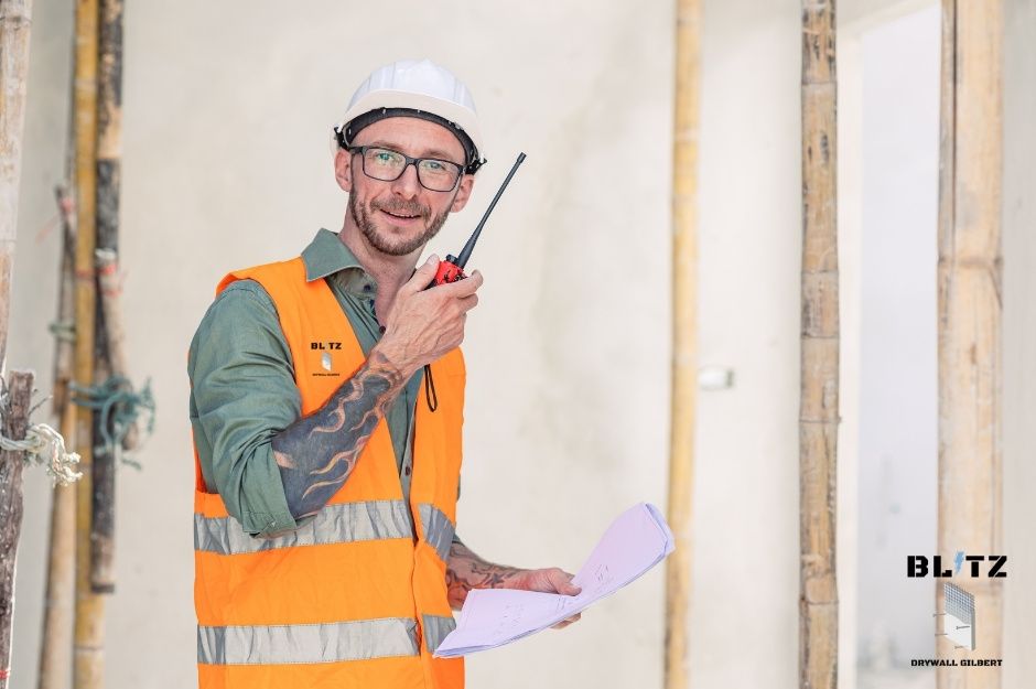 A drywall repair company worker wearing an orange high-visibility safety vest, and a white hard hat, holding blueprints or residential construction plan, while standing inside framed building structure. He has short hair and is smiling brightly near Pinnacle Pl, and S River Ridge Rd, Black Canyon, AZ.
