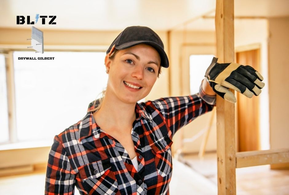 A smiling drywall repair company worker, in a red plaid shirt, stands with her hands positioned near exposed wood framing in an unfinished room with bright windows behind her at a residential construction site near E Villa Cassandra Way, and N Sunset Trail, Carefree, AZ.