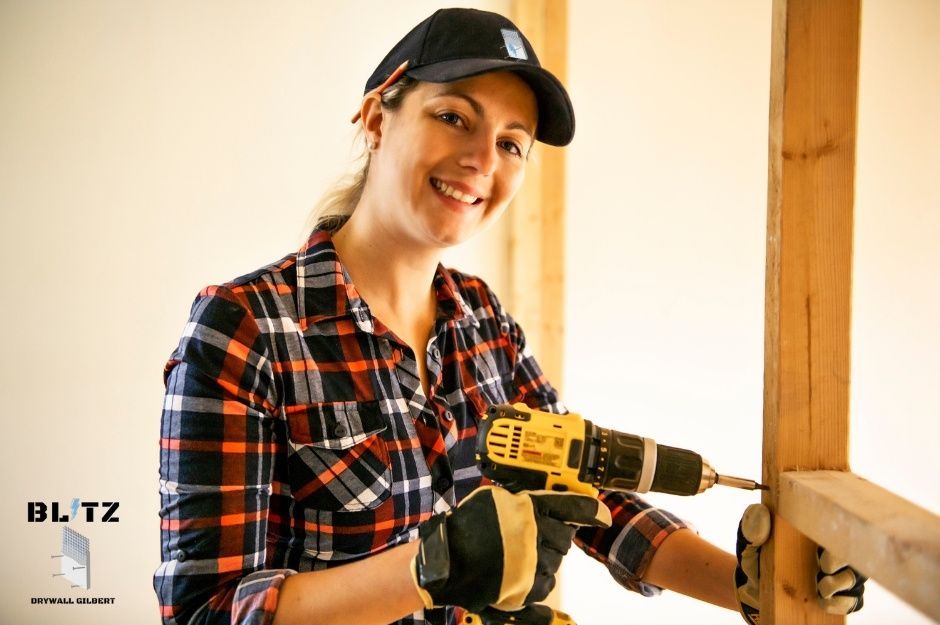A smiling drywall repair company worker wearing a black cap and plaid shirt holds a yellow cordless drill while working on wooden framing in a commercial construction site with exposed wood beams visible. She is doing a project near W Ginger Ave, and 46th St, Coolidge, AZ.