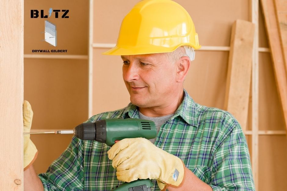 A smiling drywall repair company worker wearing a yellow hard hat, green plaid shirt, and work gloves holds a green power drill while working on a commercial drywall project with wooden studs and drywall sheets visible in background near W Toluca Dr, and N Valley Rd, Eloy, AZ.