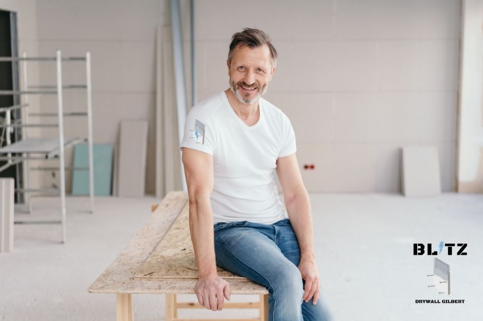 A drywall repair company professional in a white t-shirt and jeans sits on a wooden workbench in a commercial construction space. The workspace shows ongoing commercial renovation with professional lighting near E Meadow Dr, and S Country Rd in Mayer, AZ.