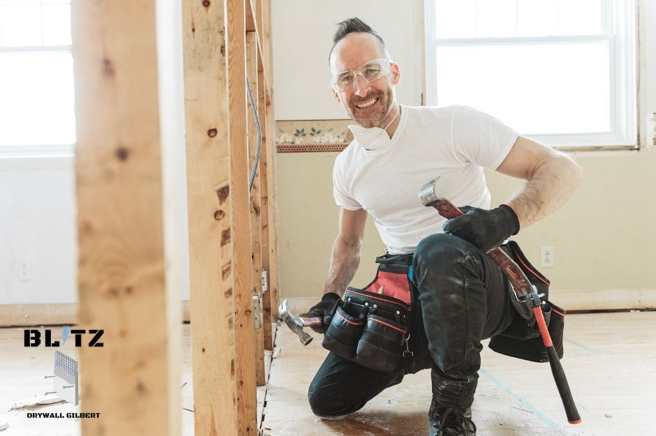 A smiling worker from a drywall repair company wearing a white t-shirt and black pants kneels while holding a hammer in an unfinished commercial area with wooden framing and windows visible. He is working at Tatum Canyon in Paradise Valley, AZ.